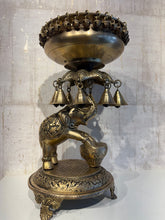 A detailed brass Urli (decorative floating bowl) with a prominent elephant base and numerous small brass bells hanging from the rim, set against a light background.